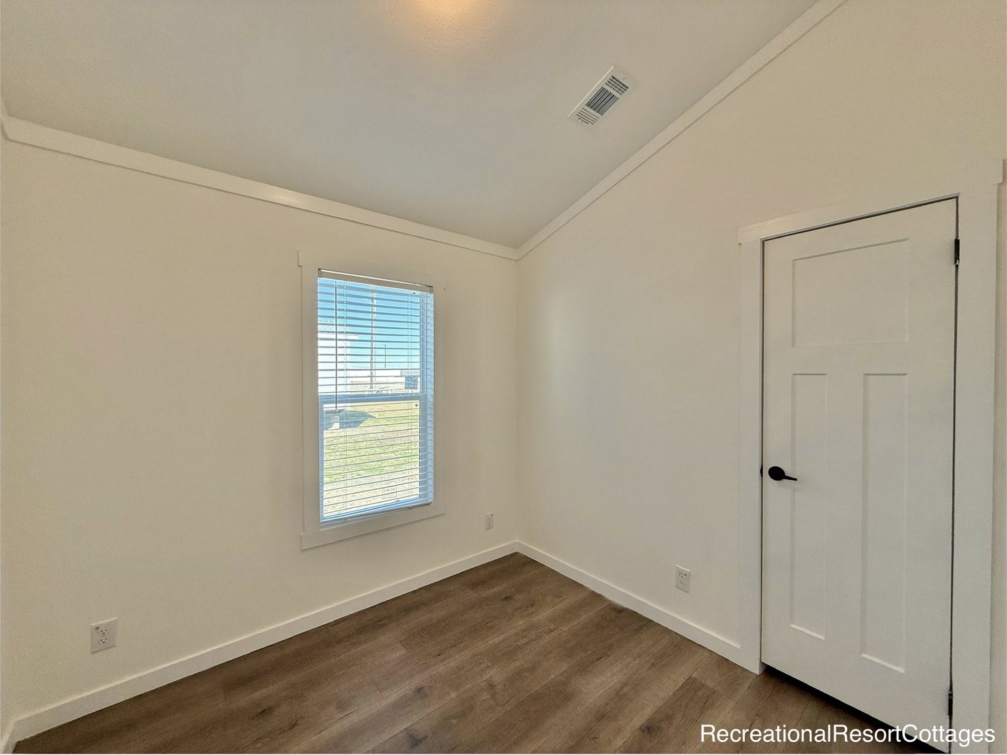 A small, bright room with cream walls, wood flooring, a window with blinds, and a closed white door on the right, conveying a minimalist feel.