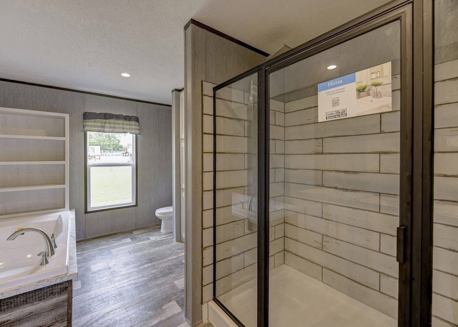 Modern bathroom with wood flooring, tiled shower enclosed by glass door, white bathtub with silver faucet, and window with dark blinds. Simple and clean design.