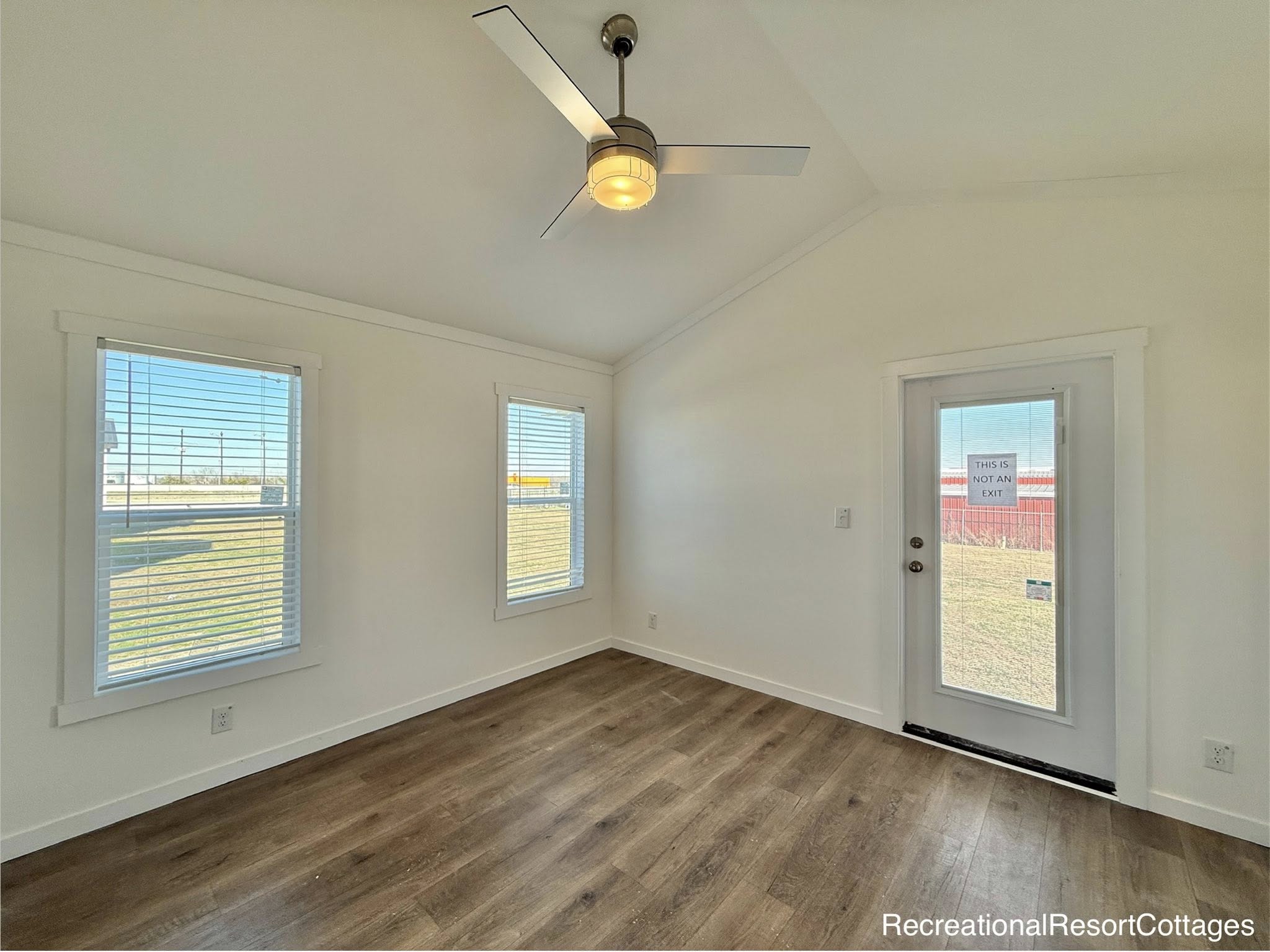 Bright, empty room with white walls, two windows, hardwood floor, and a glass door. A ceiling fan with a light hangs centrally, creating an airy feel.