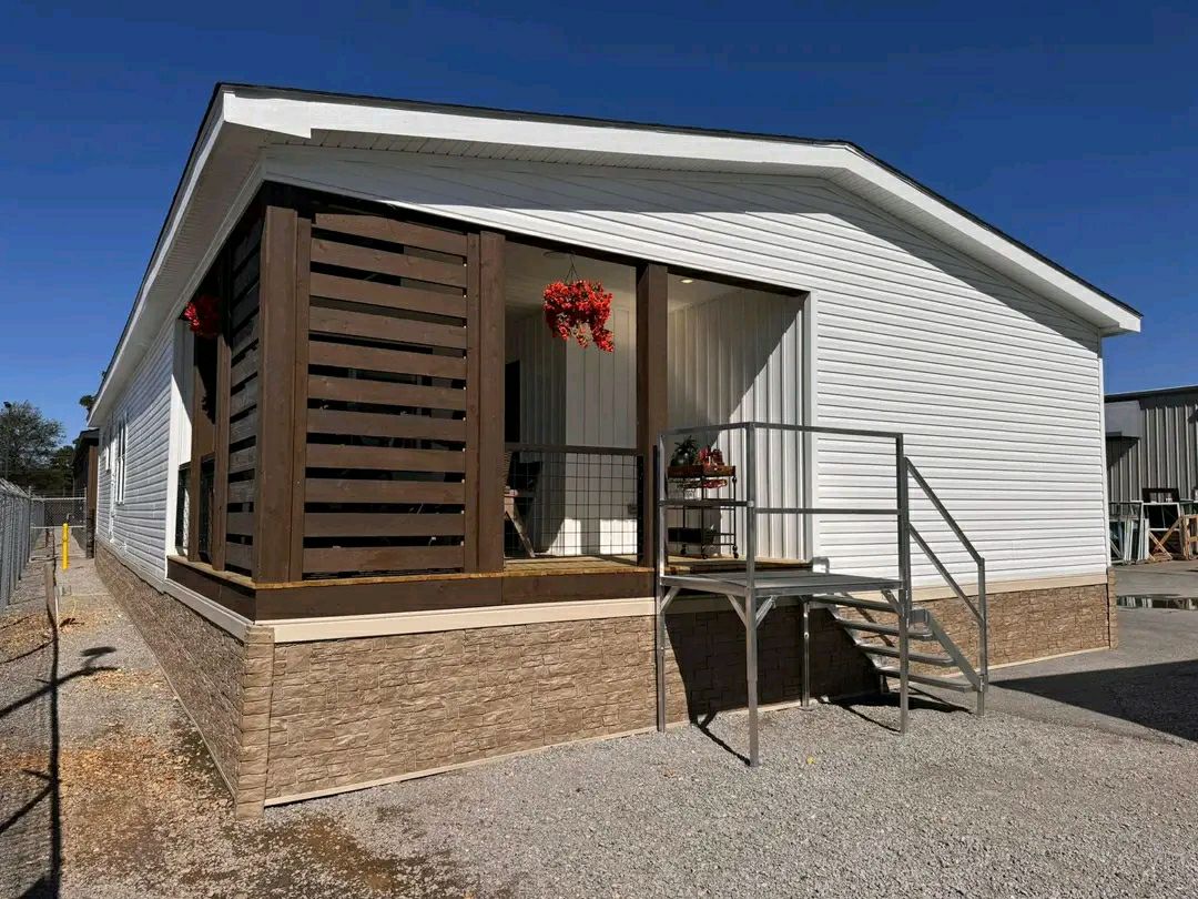 A white manufactured home with a stone foundation, set against a clear blue sky. It features brown wooden accents, stairs, and hanging red flowers, creating a cozy, inviting feel.