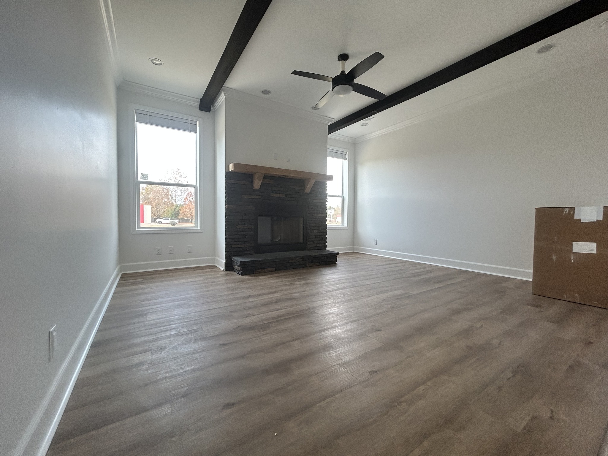 Modern living room with wooden floor, white walls, and a stone fireplace. Black ceiling beams and fan add contrast, while large windows offer natural light.