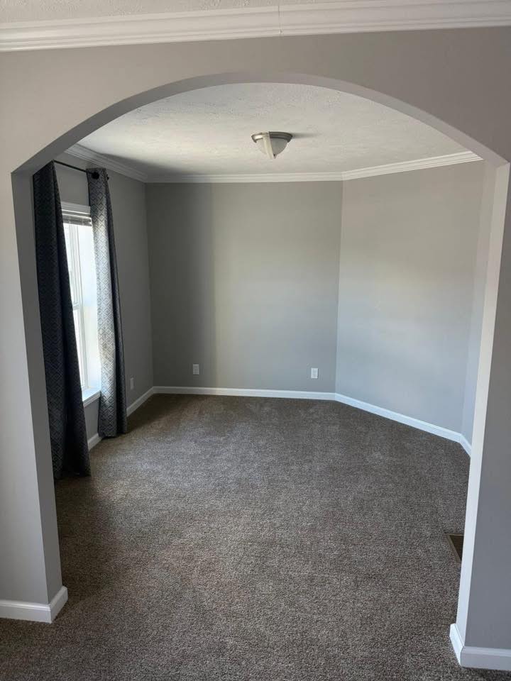 An empty room with light gray walls, an arched entryway, and brown carpet. A small window with dark curtains lets in natural light, creating a serene ambiance.