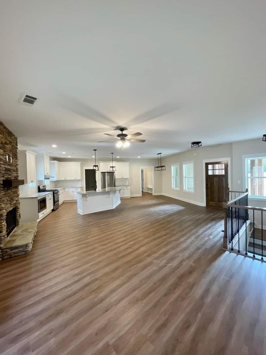 Spacious open-concept room with hardwood floors, featuring a modern kitchen with white cabinets, stone fireplace, ceiling fan, and a wooden door.
