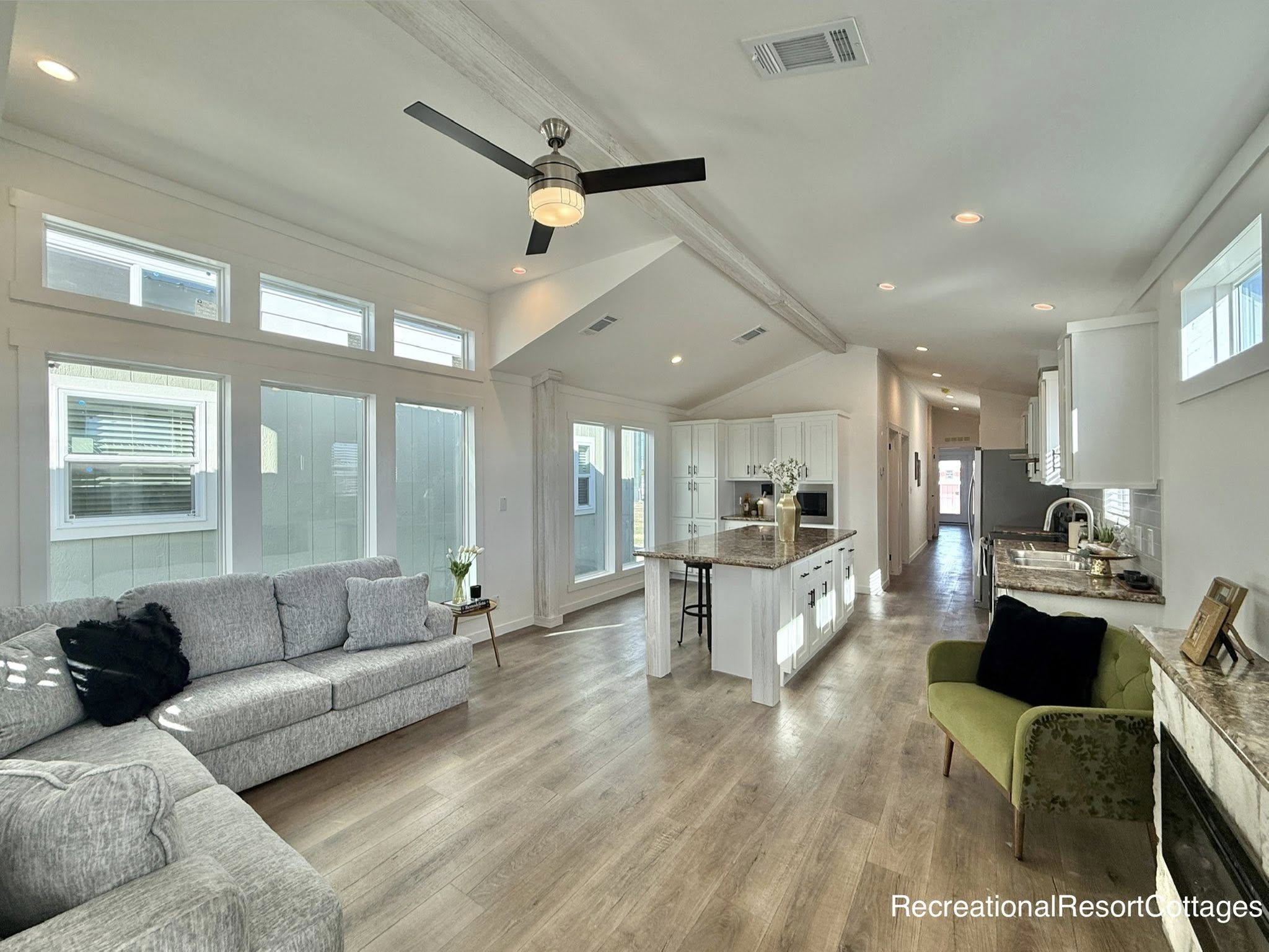 Spacious living area with vaulted ceiling, gray sectional sofa, and modern kitchen. Bright, inviting space with large windows and wood flooring.