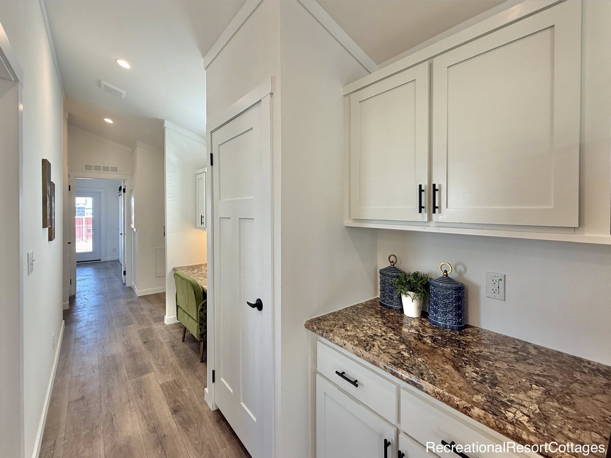 Narrow hallway with wood flooring and white cabinets on the right. Granite countertop with blue jars and plant. Bright light at the end of the corridor. Calm and tidy atmosphere.