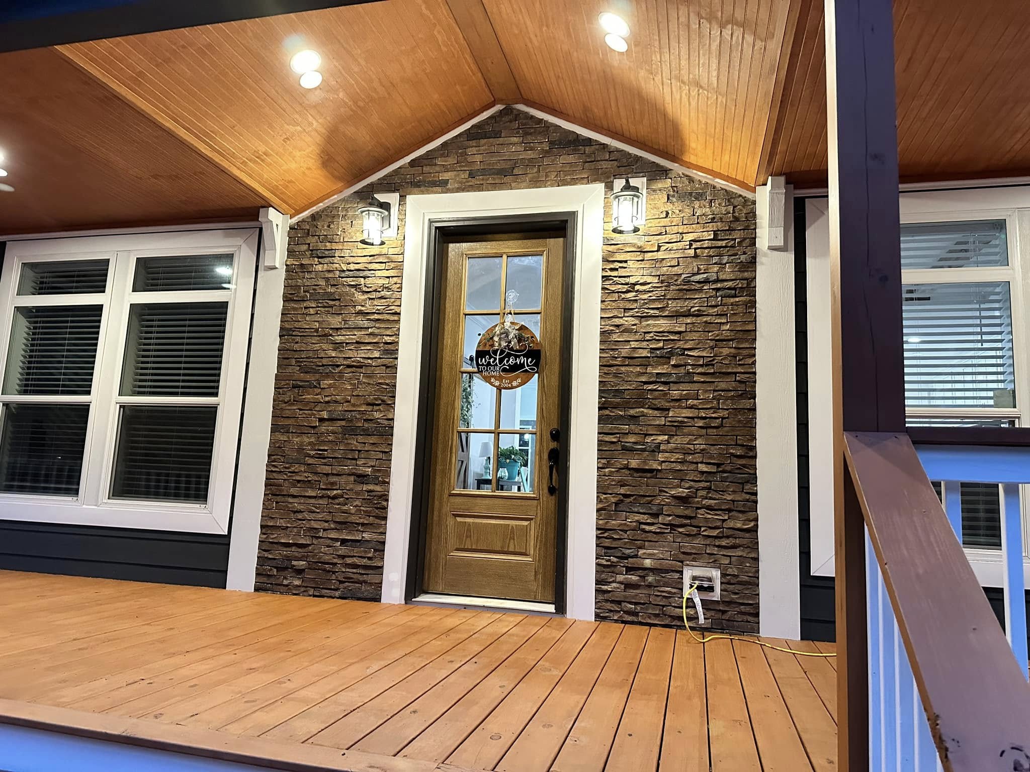 Welcoming porch with warm lighting, featuring a wooden door with a "welcome" sign, stone facade, large windows, and a polished wooden floor.