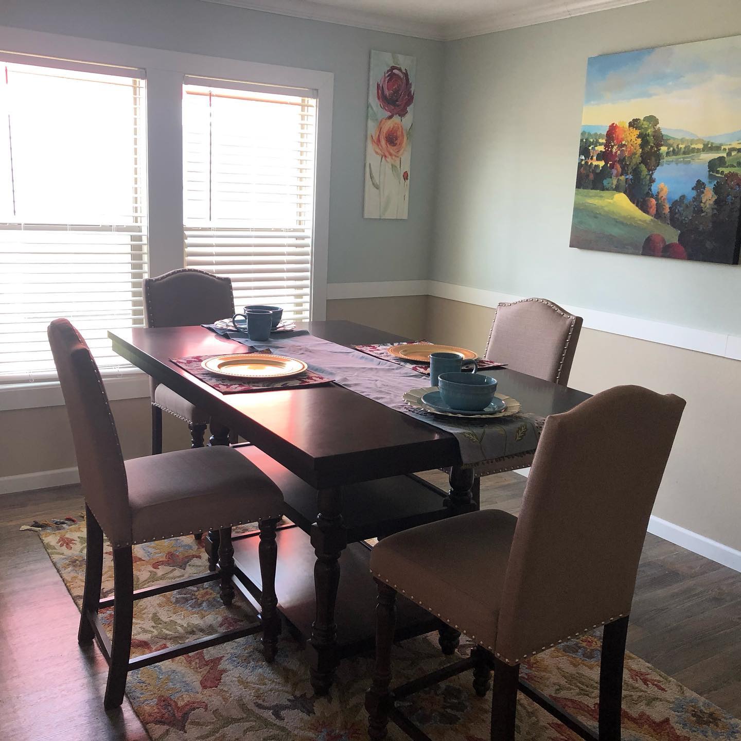 Dining room with a dark wooden table set for four, beige chairs, and blue dishware. Sunlight streams through blinds, softening the cozy atmosphere.