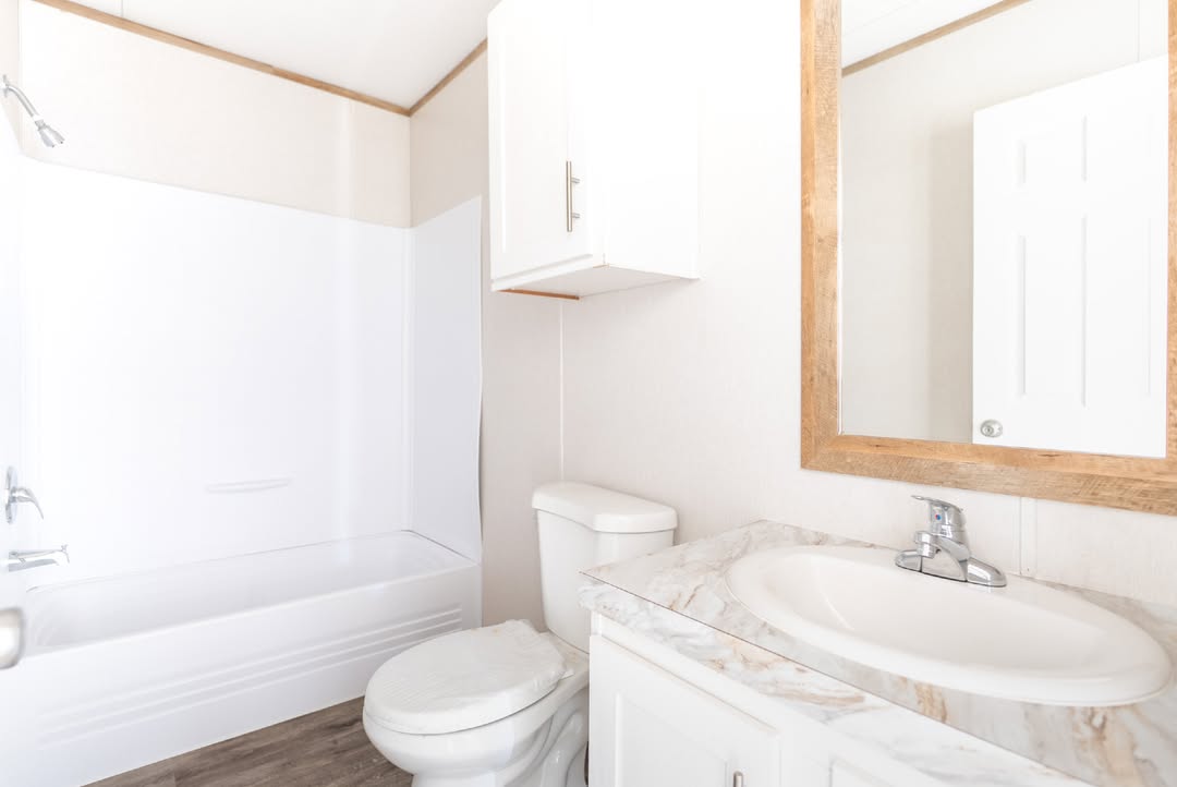 Bright, minimalist bathroom with white bathtub, a sink on a marble countertop, wooden framed mirror, white cabinets, and a toilet on hardwood floor.