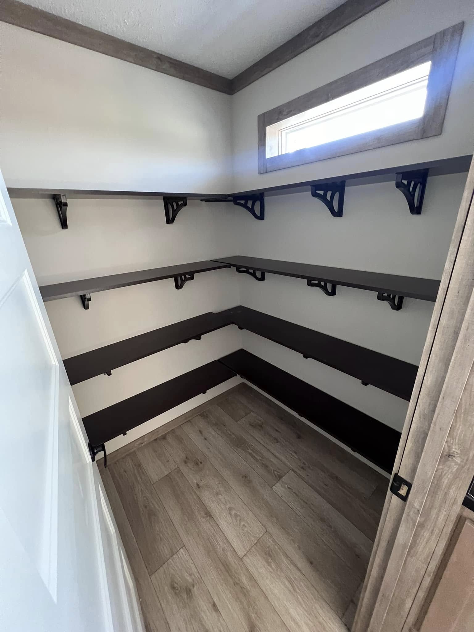 Small, empty storage room with light wood flooring, off-white walls, and dark brown corner shelving. A narrow window near the ceiling lets in natural light.