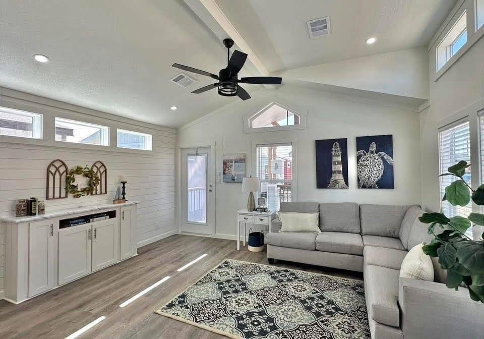 A cozy living room with a vaulted ceiling, light gray sectional sofa, elegant black ceiling fan, decorative rug, and white shiplap walls. Bright and inviting.
