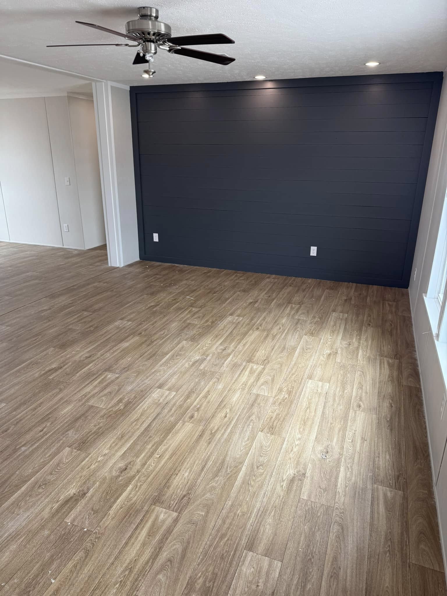 A modern, empty room with light wood laminate flooring and a dark accent wall illuminated by ceiling lights. A ceiling fan is visible, creating a calm, spacious feel.