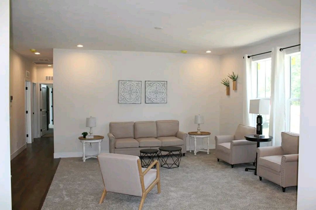 A cozy living room with beige sofas and armchairs, coffee tables, and elegant lamps on a plush carpet. Wall art and potted plants add a serene touch.