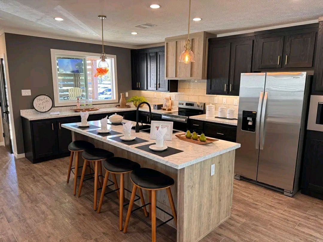 Modern kitchen with wooden floors, a central island with black stools, and pendant lighting. Dark cabinets contrast with stainless steel appliances. Cozy and inviting.