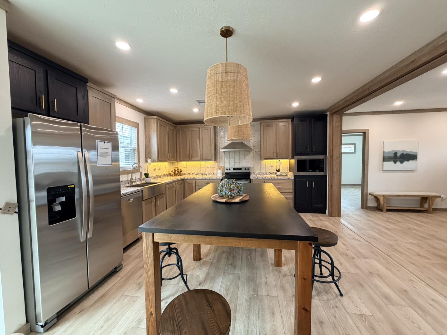 Modern kitchen with light wood cabinets, stainless steel fridge, and central island with dark wood top. Warm lighting and woven pendant lamp create a cozy ambiance.