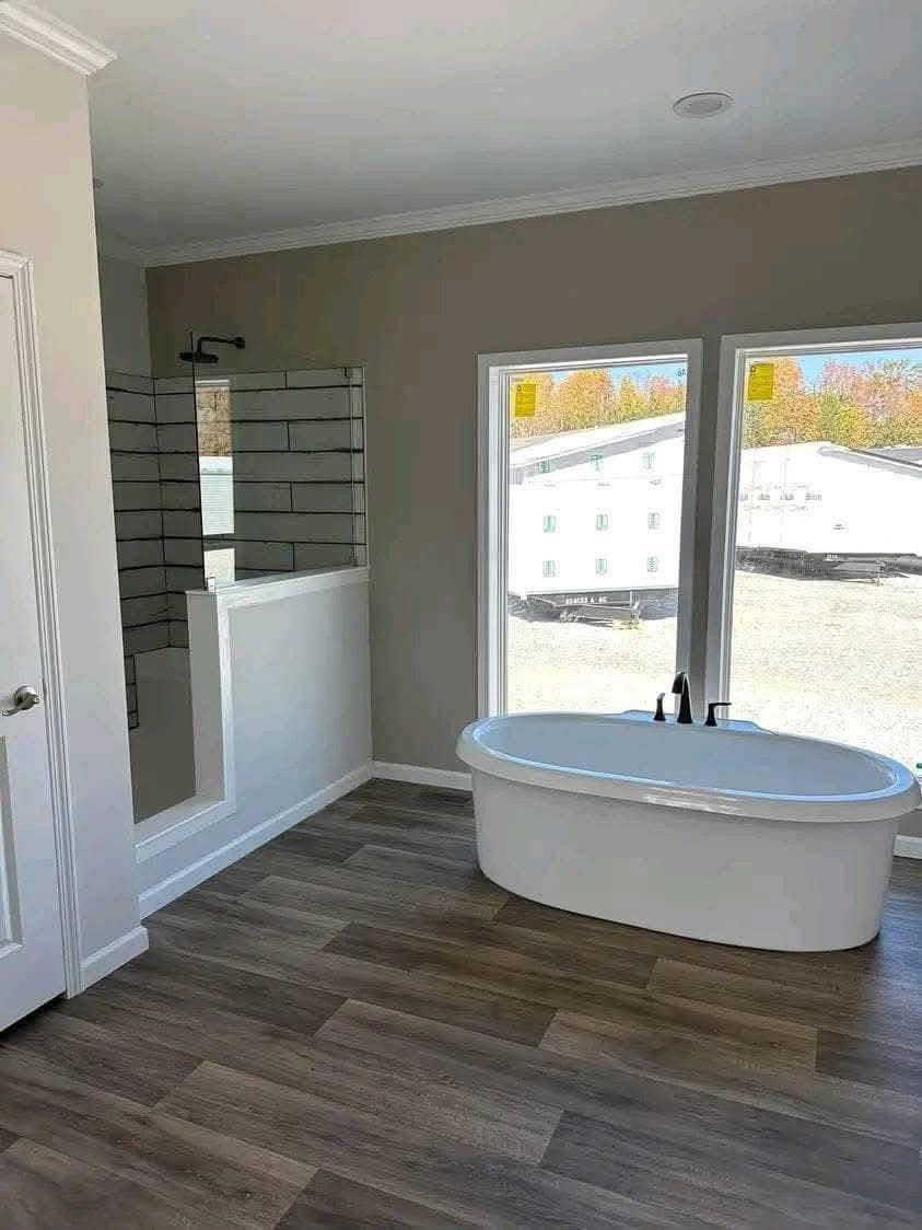 Modern bathroom with a freestanding white bathtub near large windows, wood flooring, glass-walled shower, and neutral colored walls, creating a serene feel.
