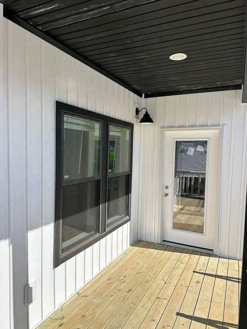 Modern porch with white paneled walls, black-framed windows, and a glass door. The wood floor and black ceiling create a sleek, welcoming feel.