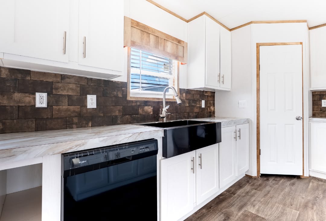 Modern kitchen with white cabinets, black sink, and marble countertops. Brown brick backsplash adds warmth. Light streams through a window above.