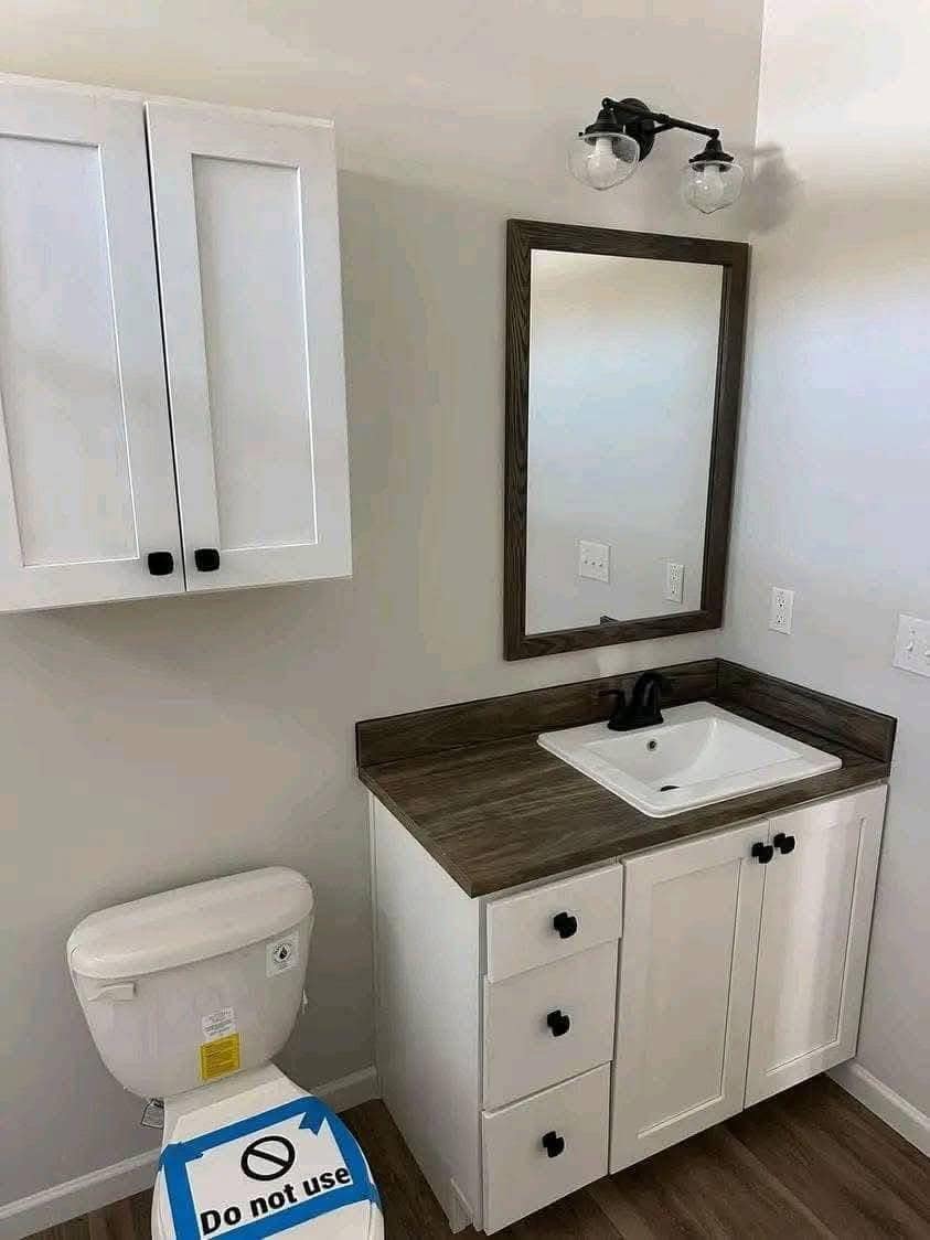 Compact bathroom with a mirror above a modern sink, wooden countertop, white cabinets, and a toilet labeled "Do not use." Bright and clean ambiance.