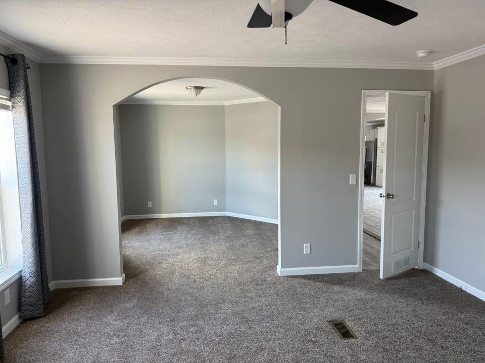 A neutral-toned living space with gray walls and carpet, featuring an arched doorway, partial view of a window with curtains, ceiling fan, and open door.