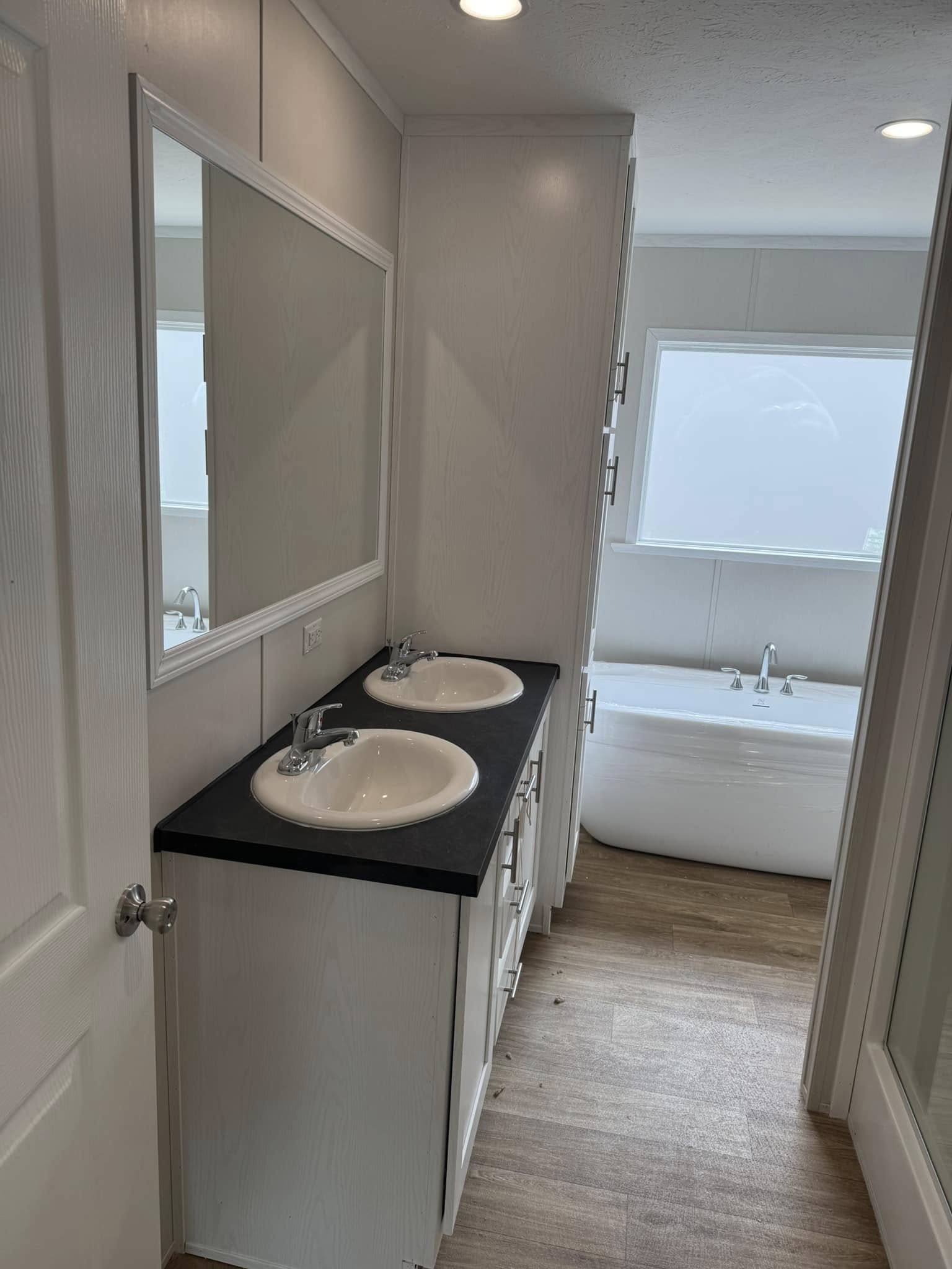 A modern bathroom with a double sink vanity, black countertop, and a large mirror. A white bathtub is visible near a frosted window, creating a bright, clean atmosphere.