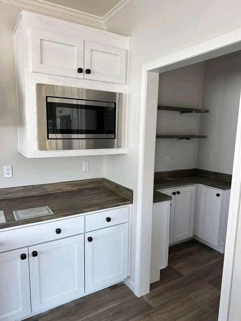 A modern kitchen with white cabinets, a built-in microwave, and wood countertops. There's an adjoining pantry area with open shelves, creating a clean, minimalist look.