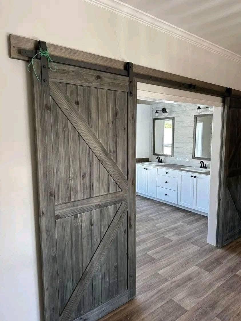 Rustic wooden barn door on a sliding rail opens to a bright bathroom with white cabinetry, dual mirrors, and wood-patterned flooring.