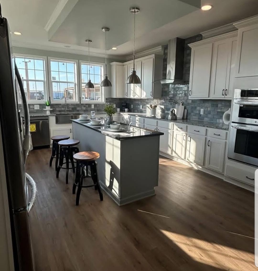 Bright kitchen with white cabinets, marble countertops, and three bar stools at the island. Pendant lights hang above; sunlight streams through large windows.