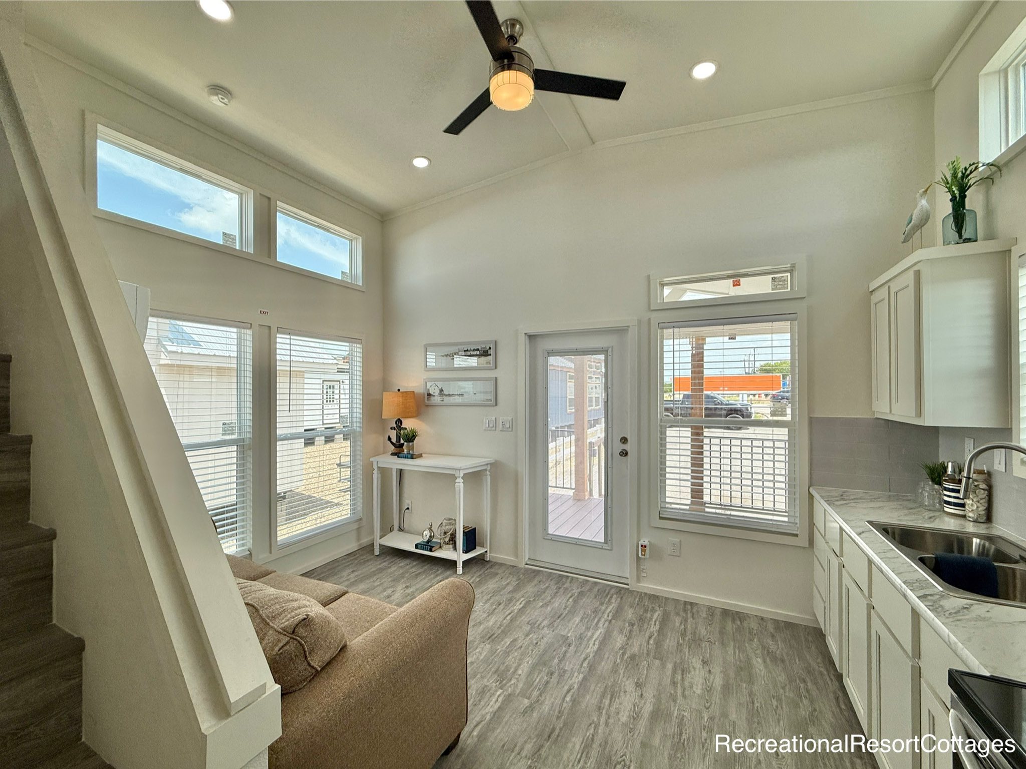Modern tiny home interior with high ceilings and abundant natural light. Features a beige sofa, white cabinets, sleek flooring, and a ceiling fan. Cozy and airy atmosphere.