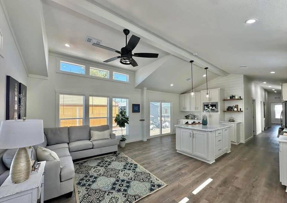 Spacious living room with gray sectional sofa, patterned rug, and modern ceiling fan. Bright kitchen features white cabinets and island, under vaulted ceiling.