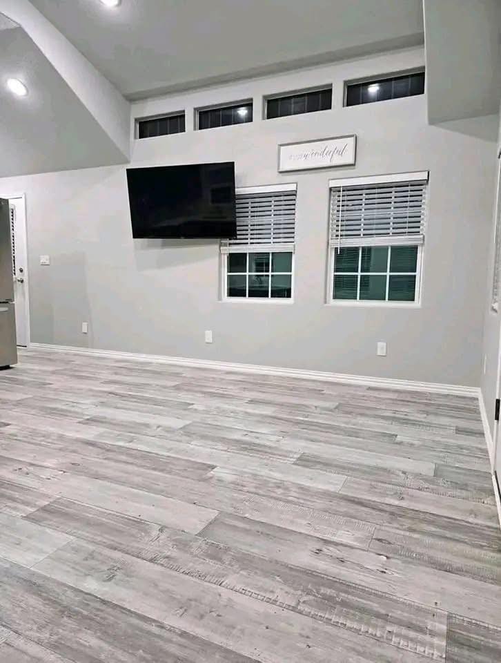 Modern, minimalist room with gray wood flooring and light gray walls. Features a wall-mounted TV, two windows with shades, and upper rectangular windows.