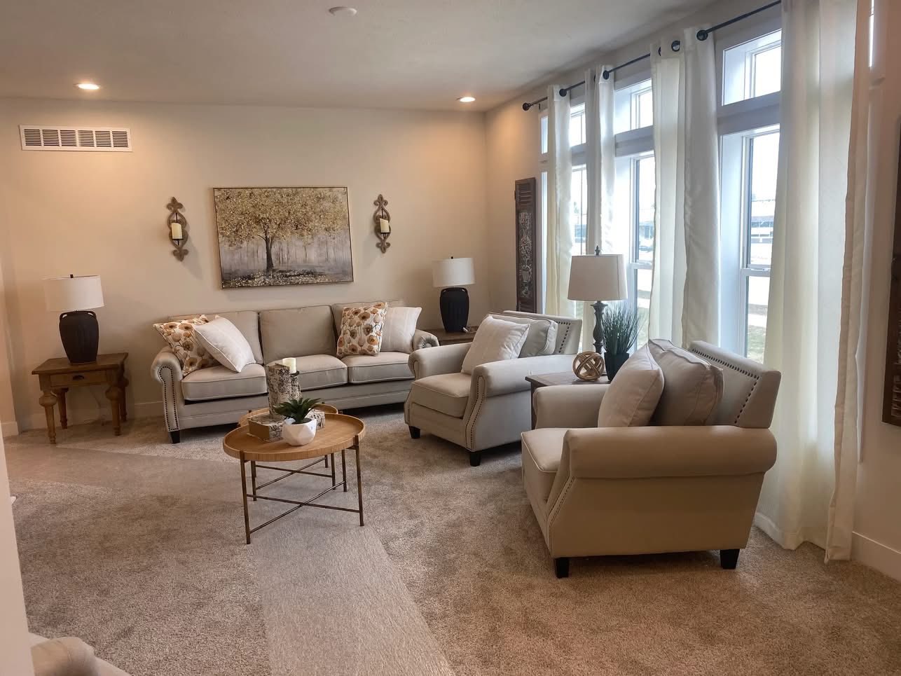 Bright living room with beige sofas and armchairs, patterned cushions, and light curtains. A round coffee table and wall art create a cozy atmosphere.