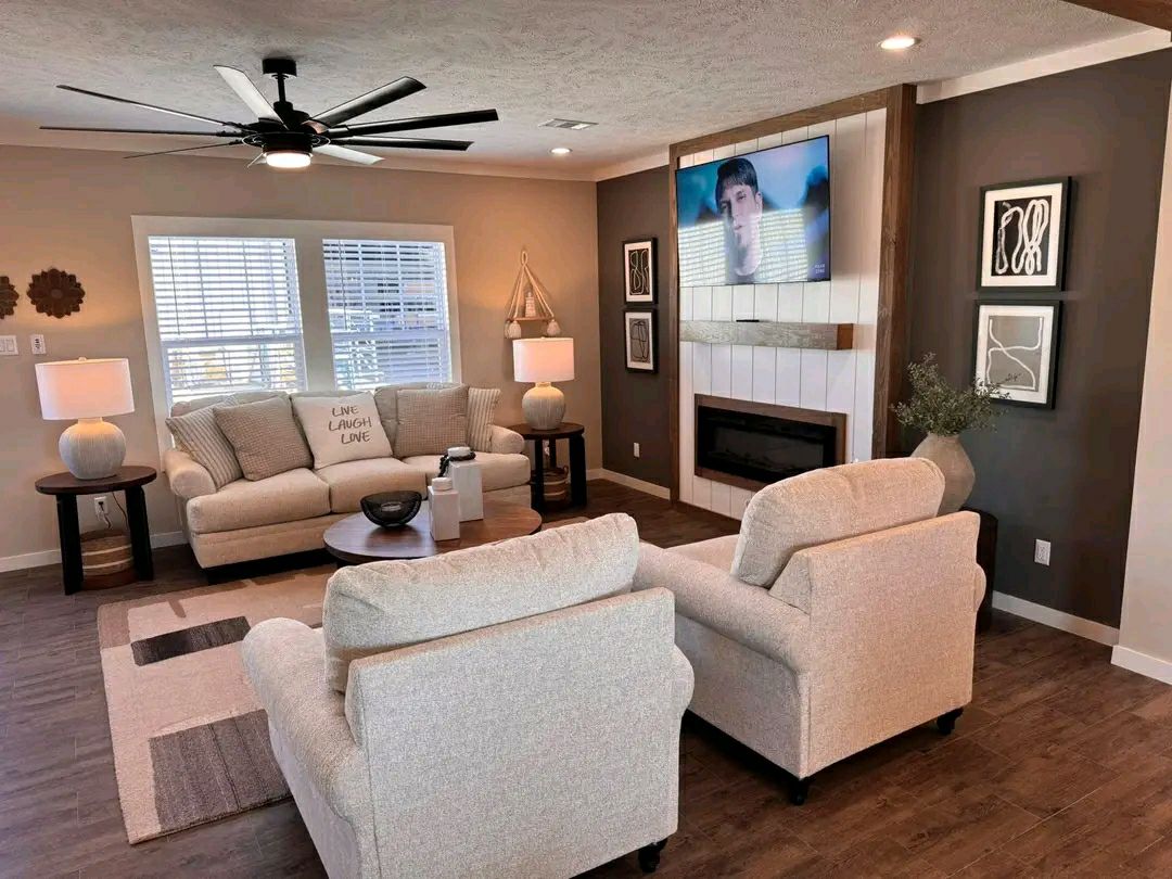 Cozy living room with plush beige sofas, modern decor, and a wall-mounted TV above a fireplace. Light streams through large windows, creating a warm ambiance.