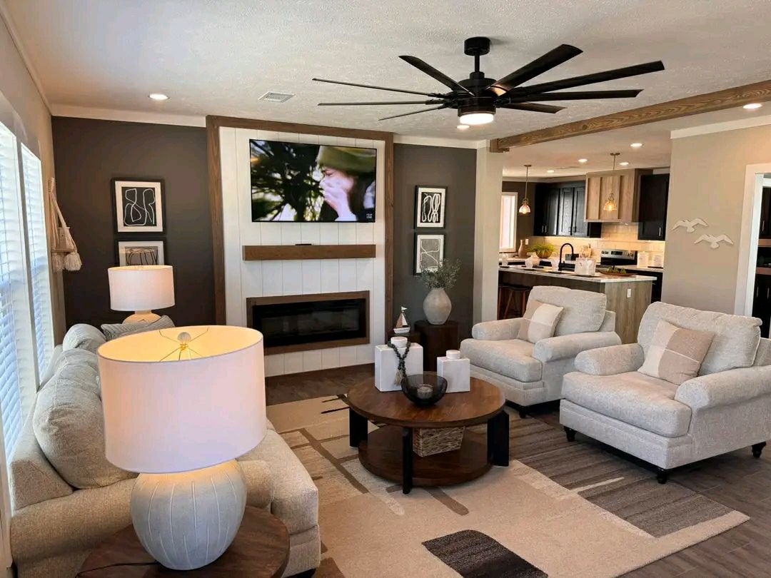 Modern living room with neutral tones, featuring two beige armchairs, a sofa, round wooden coffee table, wall-mounted TV, and ceiling fan. Cozy and inviting atmosphere.