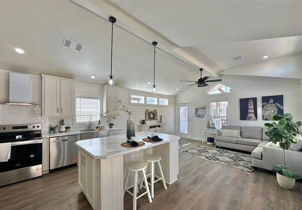 Spacious modern open-plan kitchen and living room with vaulted ceilings. The kitchen features white cabinetry, a marble island, and pendant lights. The living area has a gray sofa, plants, and wall art, creating a welcoming ambiance.