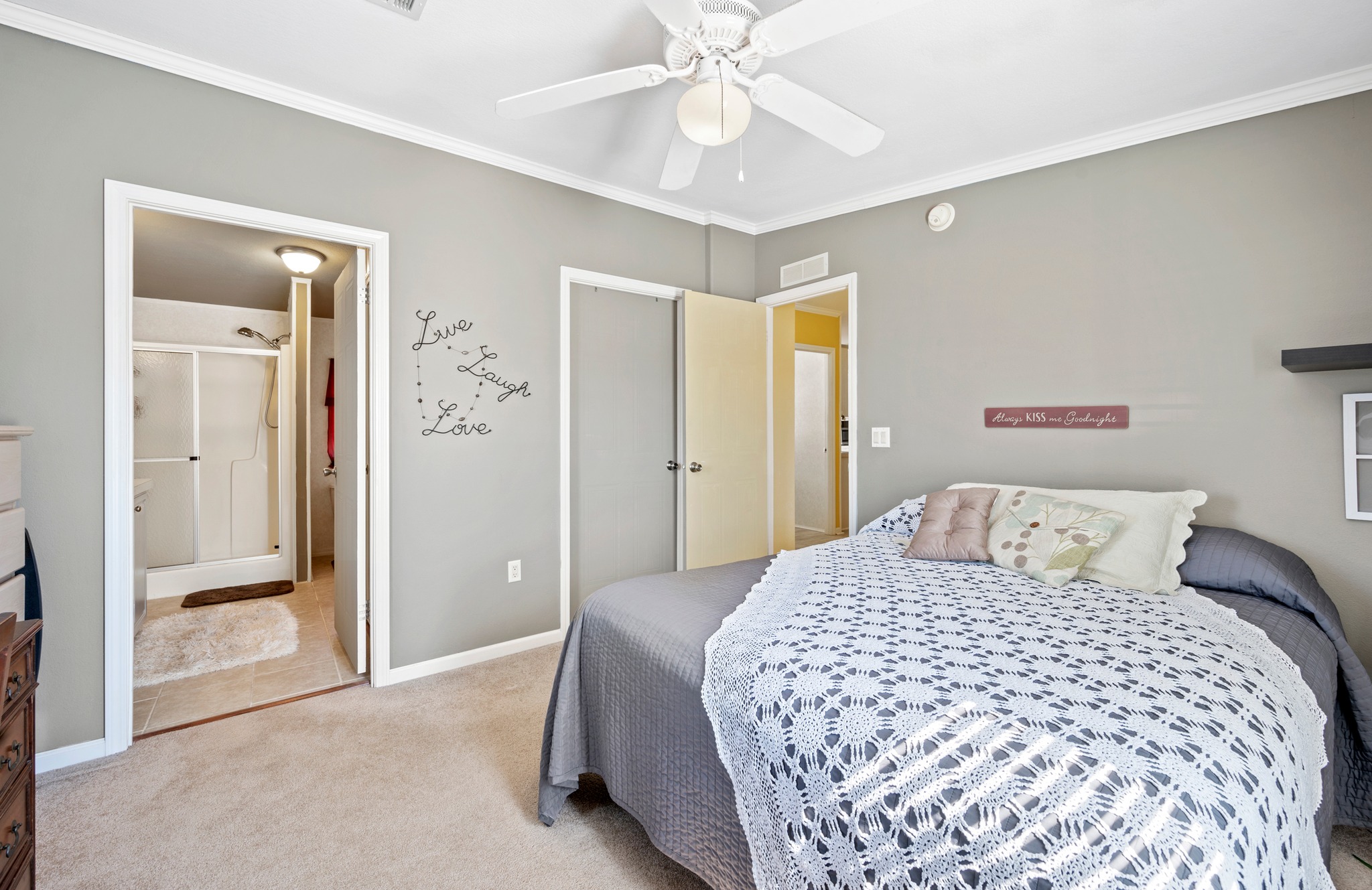 A cozy bedroom with gray walls features a neatly made bed with a crochet blanket and decorative pillows. Wall art says "Live Laugh Love." An adjacent bathroom is visible.