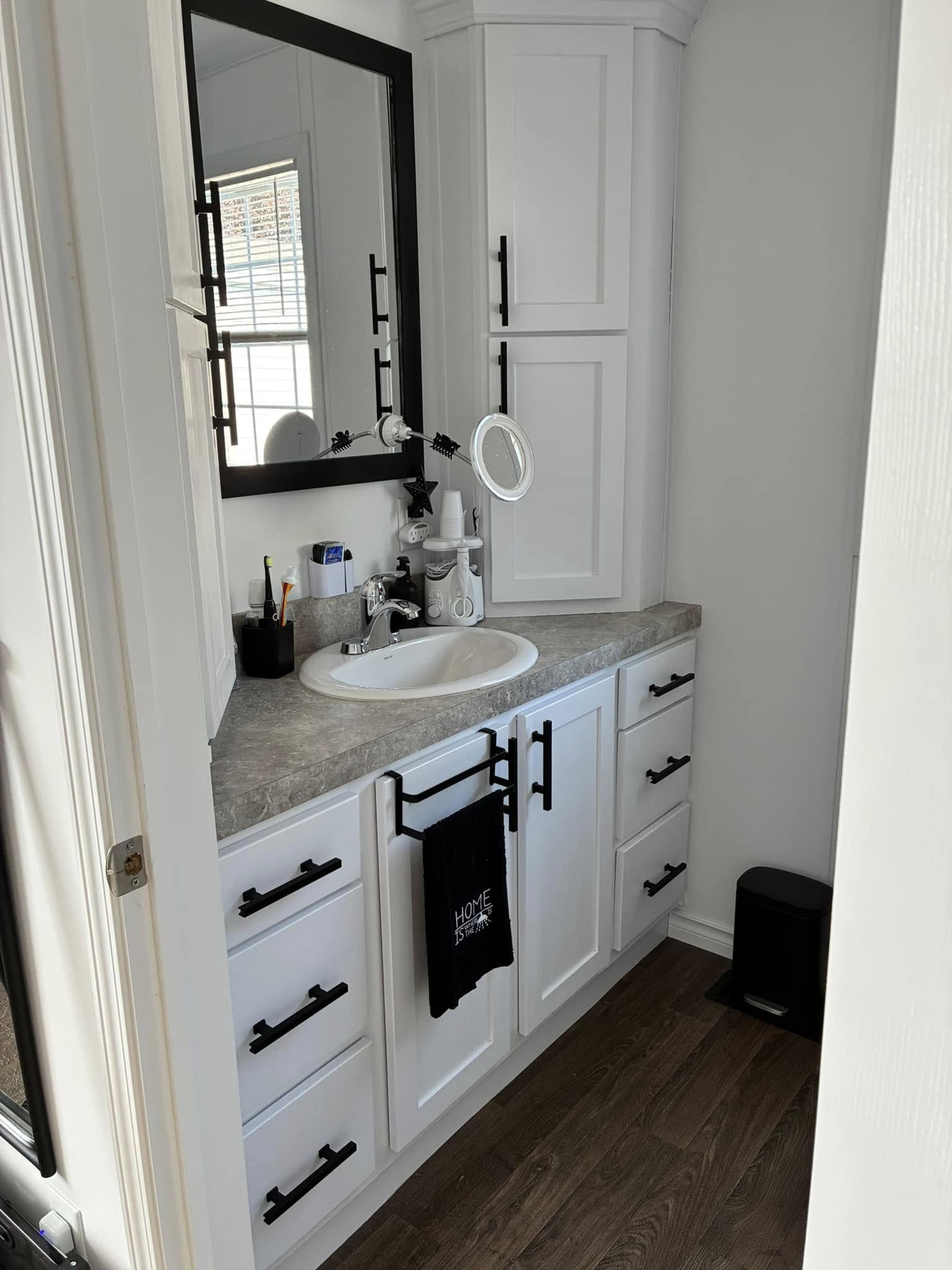 A clean bathroom corner with a white vanity, a black-framed mirror, and black hardware. A towel hangs on the rack; window light creates a cozy ambiance.