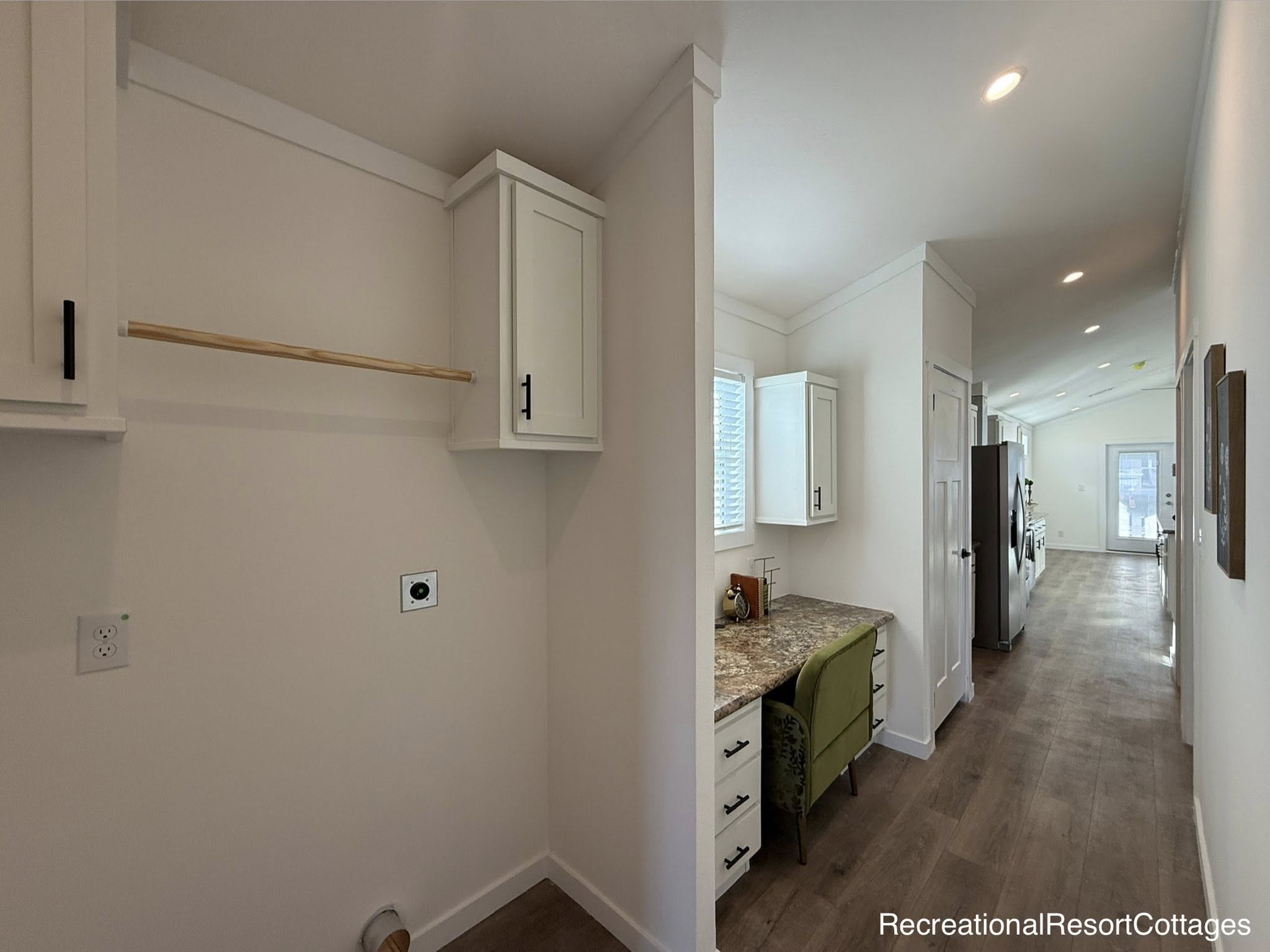 Bright modern hallway of a house with wood floors, a small desk area with a green chair, and white cabinets. A laundry area is visible on the left.