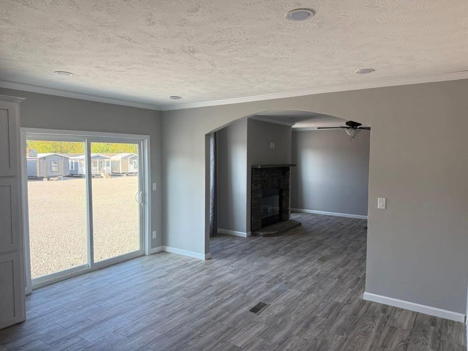 A bright, empty room with gray walls and wood-look flooring. A large sliding glass door offers a view outside. An archway leads to a cozy area with a dark fireplace. Neutral and inviting atmosphere.