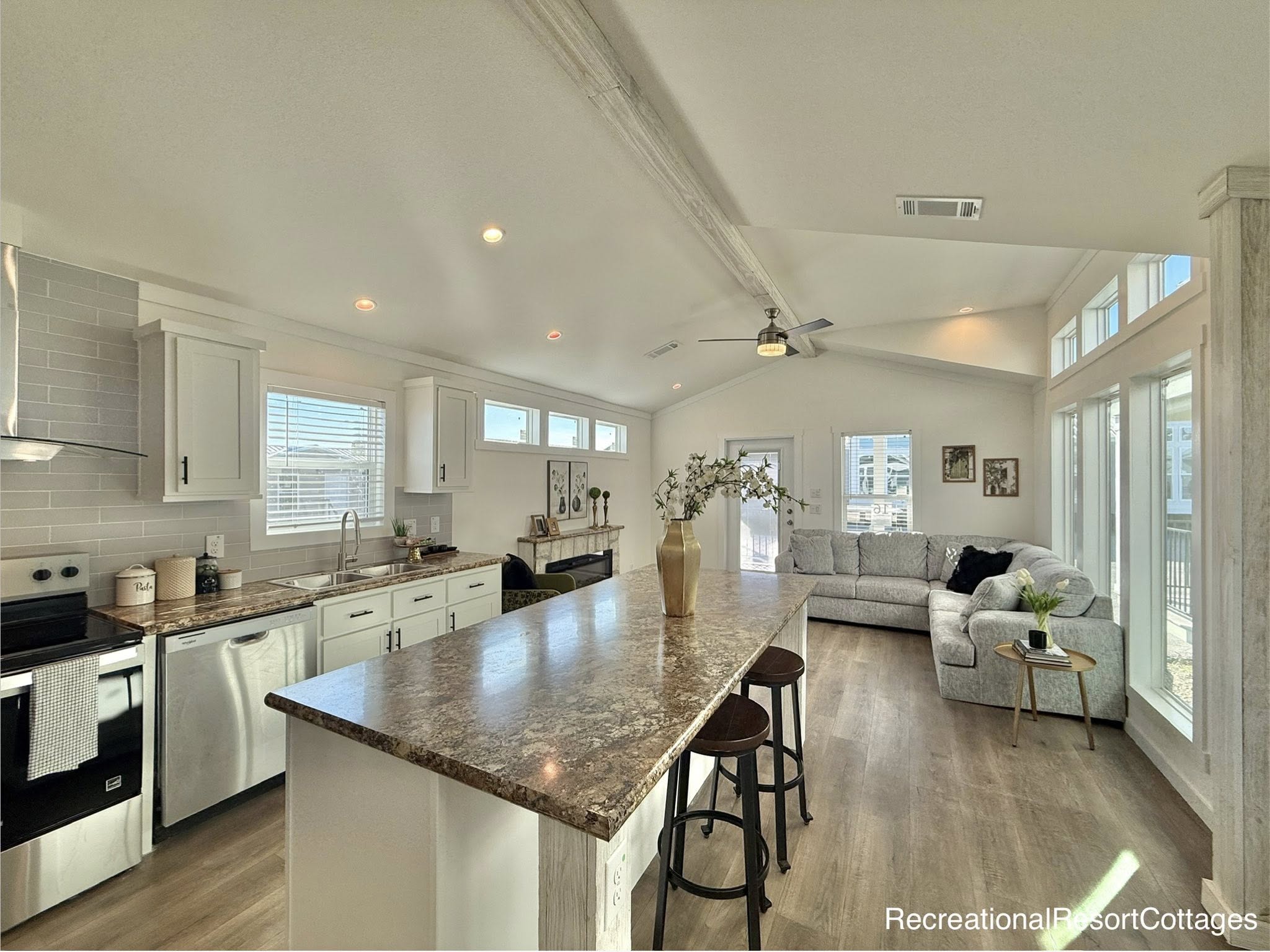 Bright open-plan kitchen and living room with high ceilings. Features a marble island with stools, cozy gray sectional, large windows, and wood flooring.