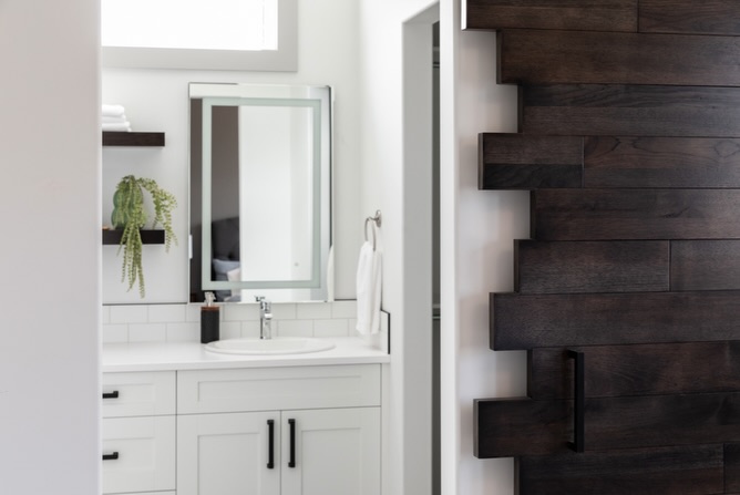 Modern bathroom with a white vanity, black handles, and a round sink. A rectangular mirror reflects part of the room. A dark wooden wall adds contrast.