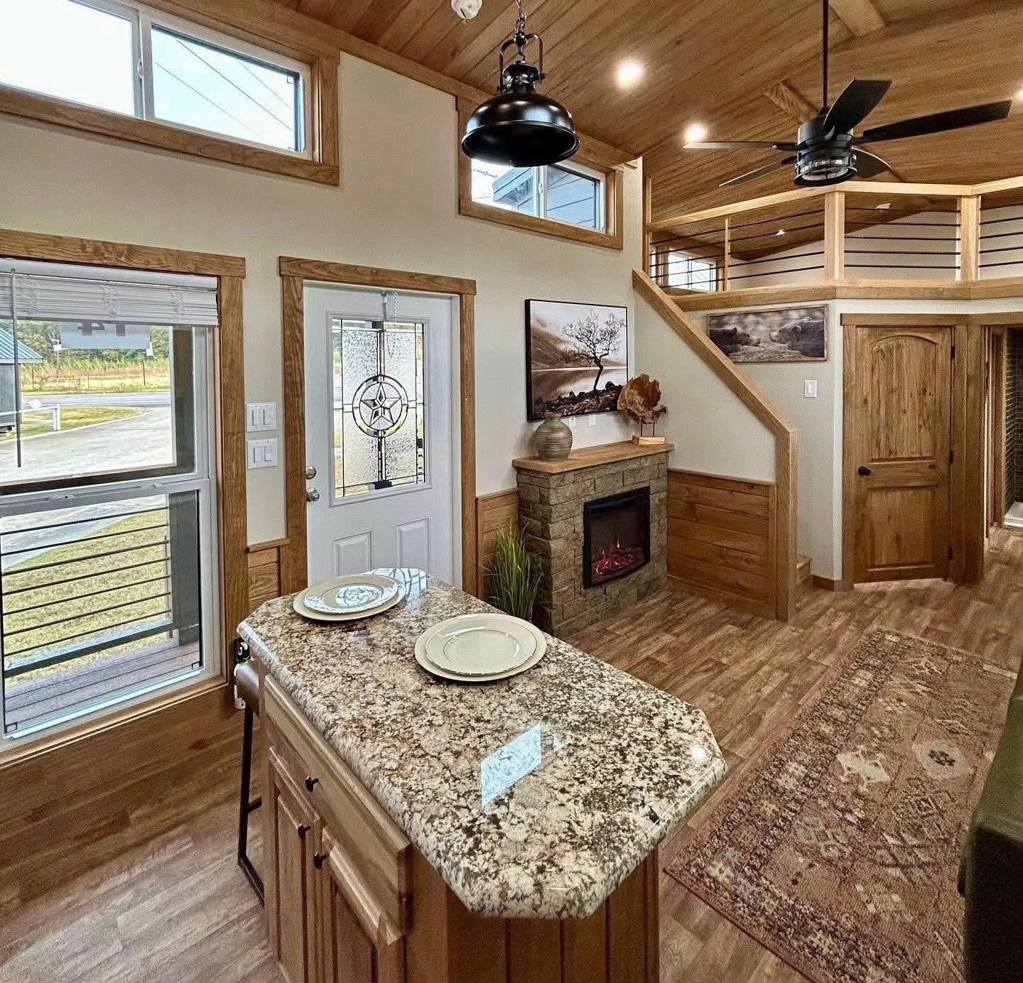 Cozy living space featuring a granite kitchen island, rustic wood accents, electric fireplace, loft area, and large windows with natural light.