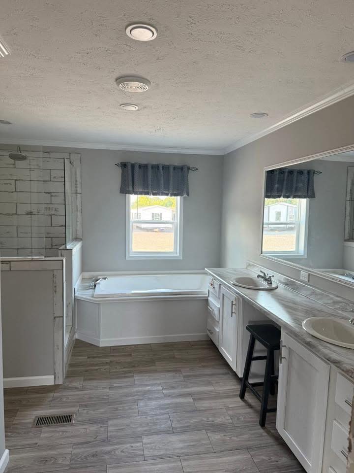 Modern bathroom with wood-look tile flooring, white cabinets, and dual sinks. A large mirror and a bathtub with a window add brightness and space.