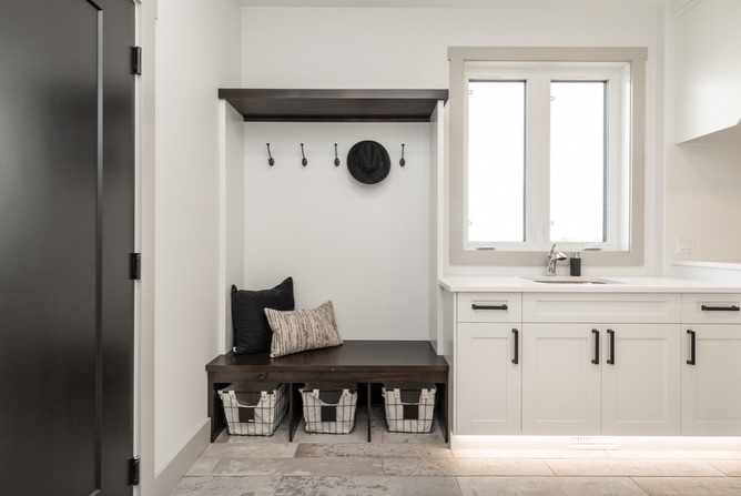 A mudroom with a dark wooden bench and coat hooks, featuring a black hat and pillows. White cabinetry with a sink and wire baskets below. Bright, clean, minimal style.