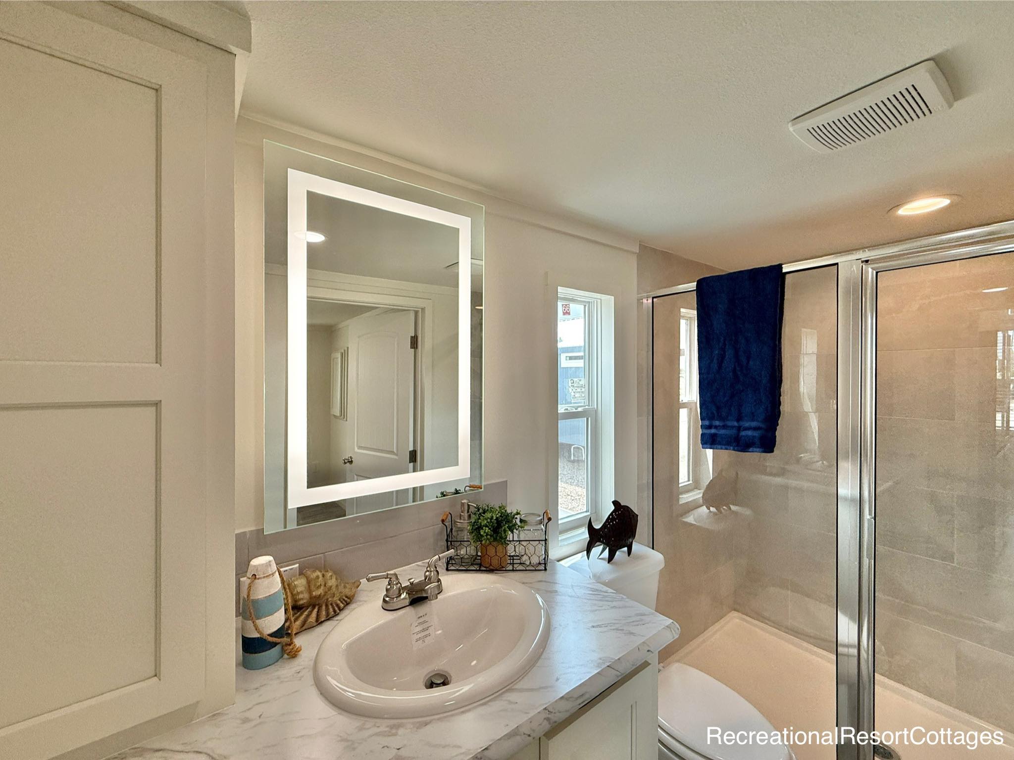 Modern bathroom with a white marble countertop and sink, illuminated mirror, glass shower, blue towel, and decorative plants, creating a fresh, cozy ambiance.