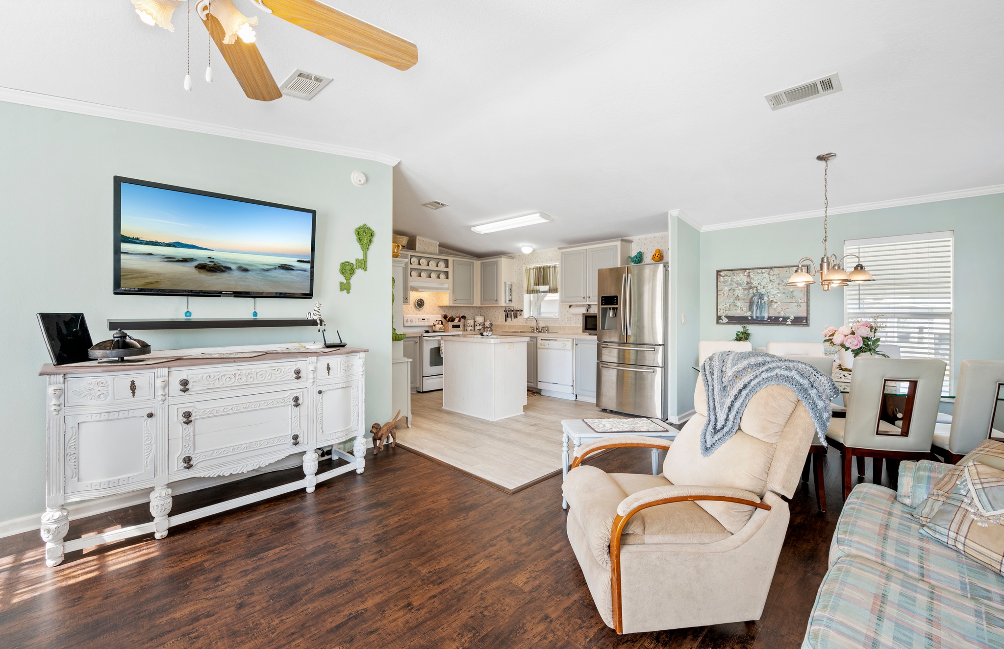 Inviting living room and kitchen open space with wooden floors, light green walls, and neutral décor. Includes a TV, armchair, and dining area. Cozy ambiance.