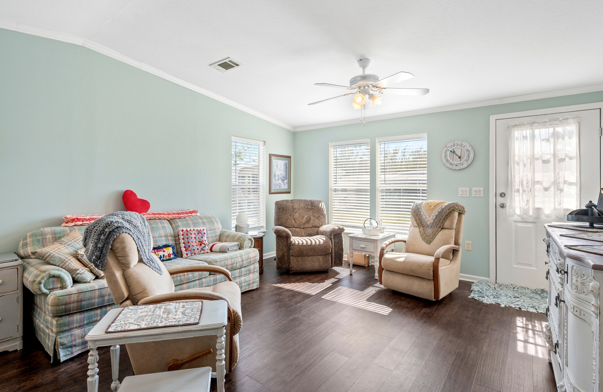 Cozy living room with soft green walls, patterned armchairs, and a plaid loveseat. Sunlight streams through windows, creating a warm, inviting atmosphere. A ceiling fan overhead and a wall clock add to the relaxed setting.