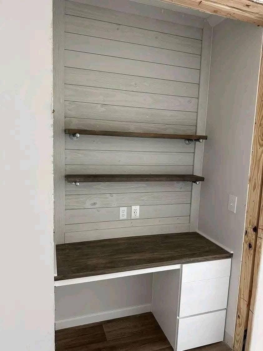 A minimalist wooden workspace features a white shiplap wall with two dark wood shelves. Below, a desk with drawers is kept clean and simple.
