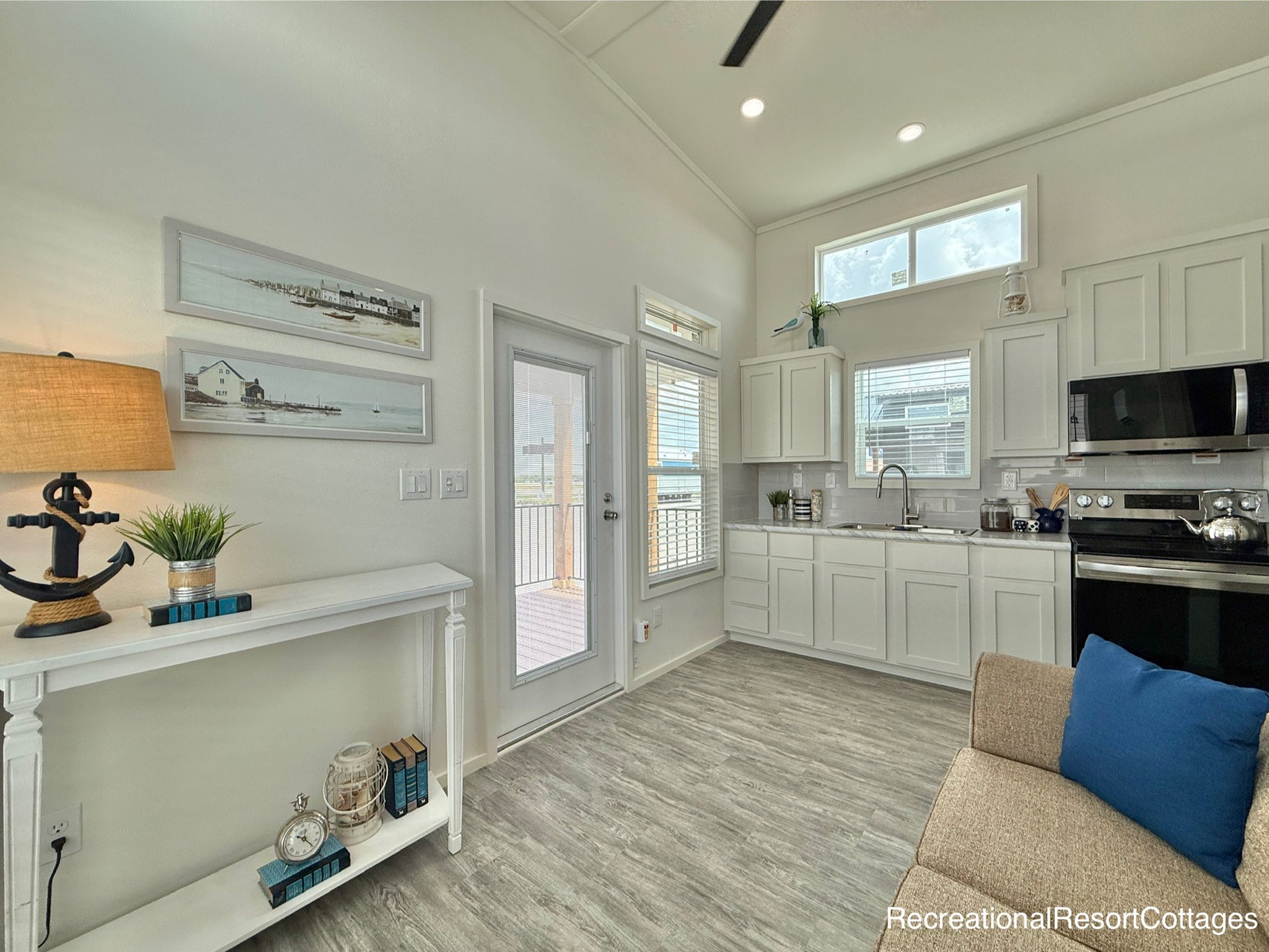 Cozy living space with white walls, light wood flooring, and a modern kitchen. Decor includes a nautical lamp, plant, and seascape paintings. Natural light pours in through large windows and a glass door.