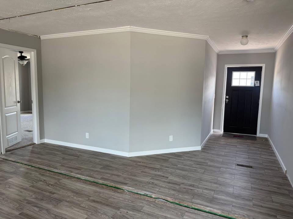 An empty room with gray walls, wood-style flooring, and a white baseboard. A black door with a window is on the right, creating a bright, clean atmosphere.