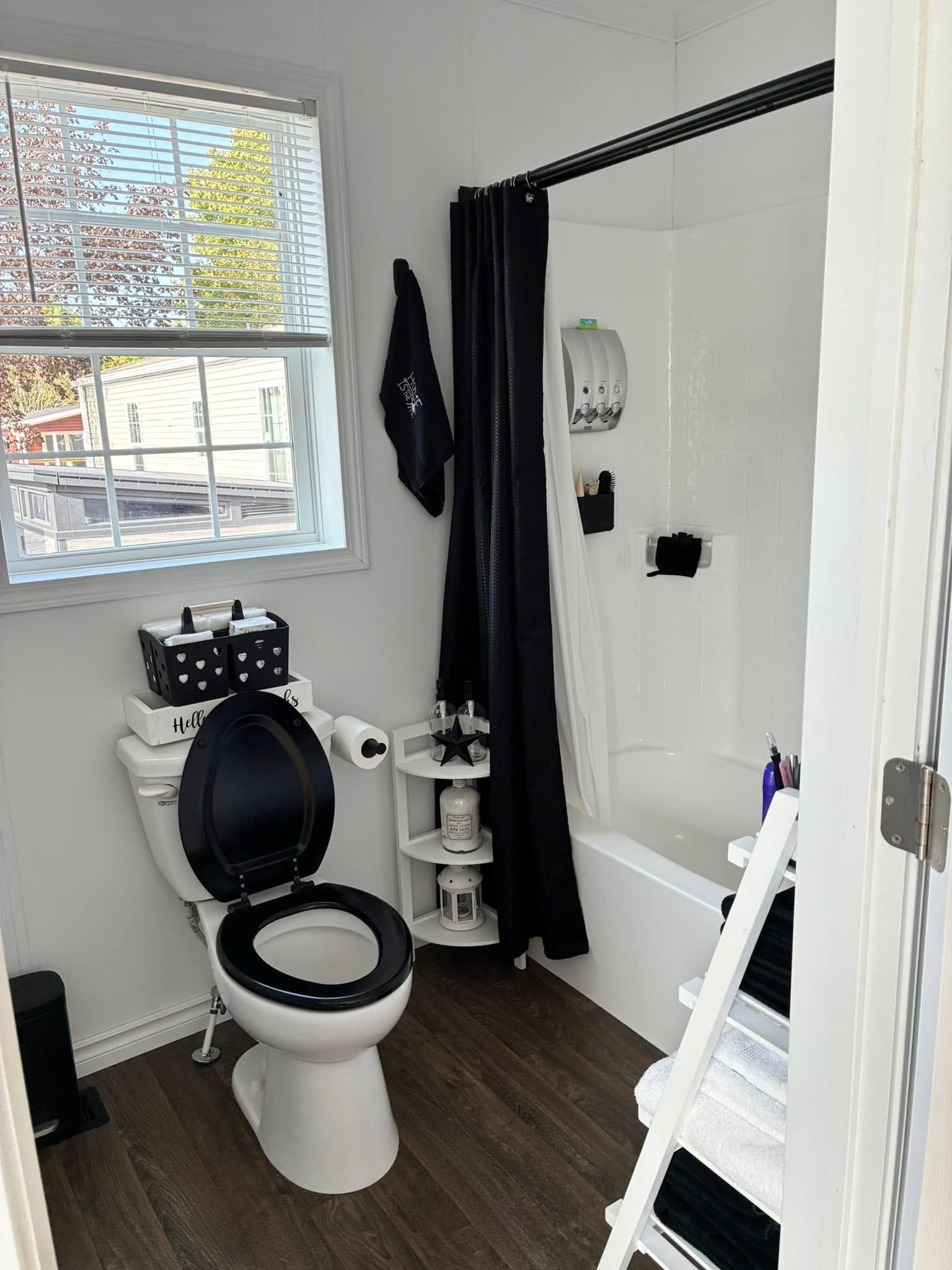 Compact bathroom with a window revealing outdoor greenery. Toilet with a black seat, nearby wooden shelf with toiletries, and a shower with dark curtains. Clean and organized.
